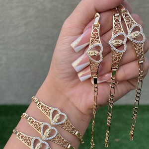 May include: Three gold bracelets with intricate heart-shaped designs featuring a Virgin Mary pendant with clear stones. The bracelets are stacked on a person's wrist.