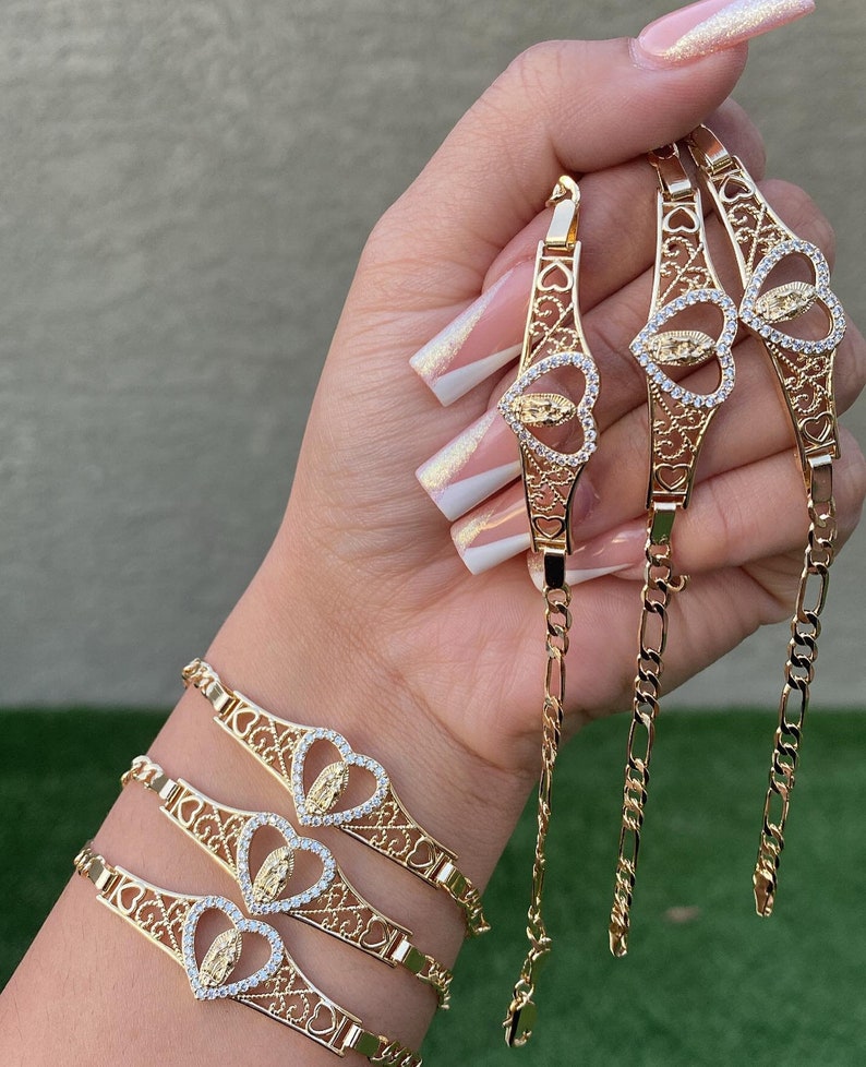 May include: Three gold bracelets with intricate heart-shaped designs featuring a Virgin Mary pendant with clear stones. The bracelets are stacked on a person's wrist.