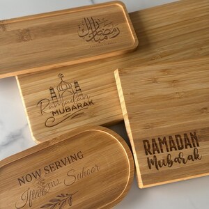 May include: Four wooden serving trays with engraved Ramadan greetings. The trays are made of natural bamboo and feature different designs, including a mosque, the Arabic phrase "Ramadan Kareem", and the text "Ramadan Mubarak".
