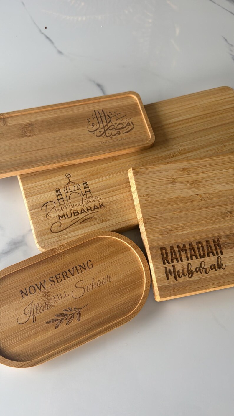 May include: Four wooden serving trays with engraved Ramadan greetings. The trays are made of natural bamboo and feature different designs, including a mosque, the Arabic phrase "Ramadan Kareem", and the text "Ramadan Mubarak".