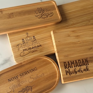 May include: Four wooden serving trays with engraved Ramadan greetings. The trays are made of light brown bamboo wood and feature different designs, including a mosque, Arabic calligraphy, and the phrase "Ramadan Mubarak".