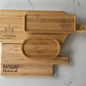 May include: Three wooden serving trays with engraved Ramadan greetings. The largest tray has a handle and the text "Ramadan Mubarak" in Arabic. The middle tray has the text "Now Serving,  Iftar & Suhoor". The smallest tray has the text "Ramadan Mubarak".