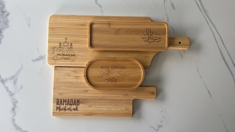 May include: Three wooden serving trays with engraved Ramadan greetings. The largest tray has a handle and the text "Ramadan Mubarak" in Arabic. The middle tray has the text "Now Serving,  Iftar & Suhoor". The smallest tray has the text "Ramadan Mubarak".