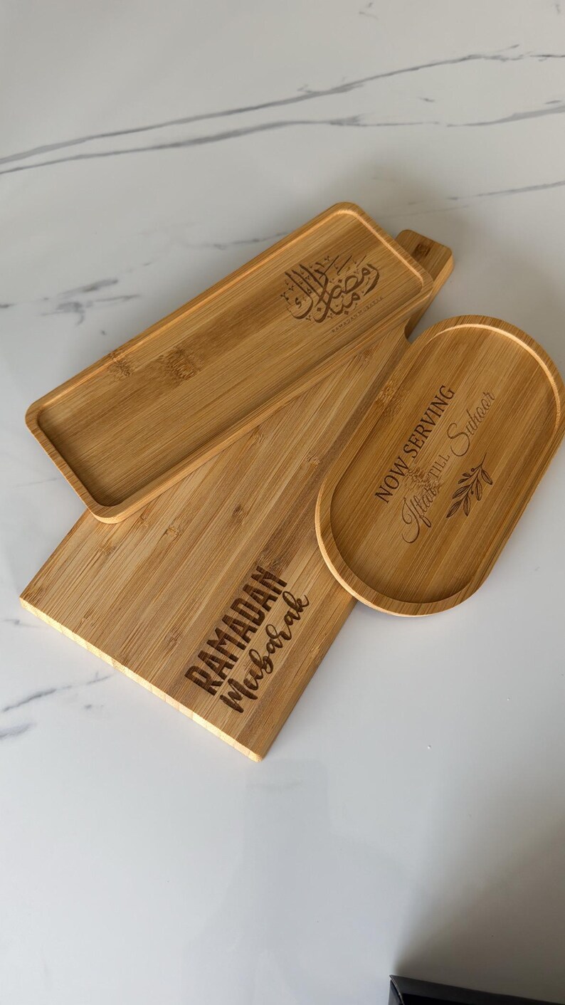 May include: Three wooden serving trays with engraved text. The largest tray says "Ramadan Mubarak". The medium tray says "Now Serving Iftar & Suhoor". The smallest tray has Arabic calligraphy.