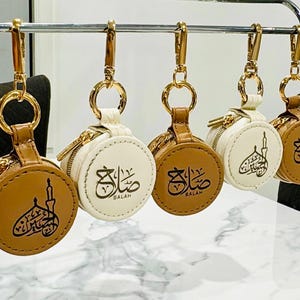 May include: Five round, zipped pouches with gold-tone lobster claw clasps and rings. The pouches alternate between tan and white colours, each featuring black Arabic calligraphy and the word SALAH printed on the front surface of the circular case.