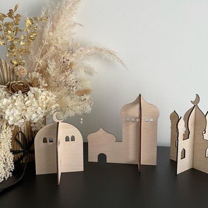 Op de afbeelding: Drie houten moskee-vormige decoraties in verschillende maten. De decoraties zijn gemaakt van lichtbruin hout en hebben ingewikkelde uitsnijdingen. De decoraties zijn gerangschikt op een zwart oppervlak.