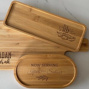 May include: Three wooden serving trays with engraved text. The largest tray is engraved with "Ramadan Mubarak". The middle tray is engraved with Arabic calligraphy. The smallest tray is oval shaped and engraved with "Now Serving After Iftar & Suhoor".