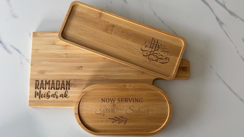 May include: Three wooden serving trays with engraved text. The largest tray is engraved with "Ramadan Mubarak". The middle tray is engraved with Arabic calligraphy. The smallest tray is oval shaped and engraved with "Now Serving After Iftar & Suhoor".