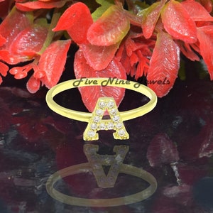 May include: A gold ring with a letter A made of small clear stones. The ring is on a black surface with red flowers in the background.