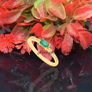 以下が含まれることがあります： 緑のエメラルドと小さな透明な石がバンドにセットされた金のリング。リングは赤い花のある黒い表面の上に置かれています。