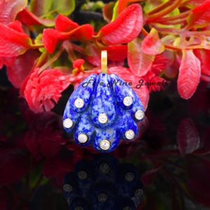 May include: A blue pendant with a gold bail, featuring a fan-like design with white accents. The pendant is set against a backdrop of red and green foliage, creating a striking contrast. The pendant's design is mirrored in its reflection.