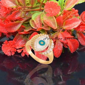 May include: Gold ring with a white and blue evil eye charm. The ring is set with clear cubic zirconia stones. The ring is on a red and green floral background.