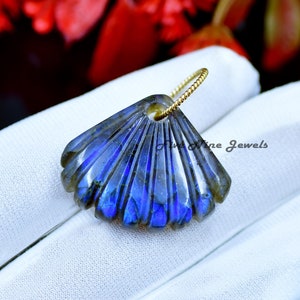 May include: A blue labradorite gemstone carved into a seashell shape, with a gold-toned chain attached. The pendant is displayed on a white surface.
