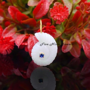 Op de afbeelding: Witte jade hanger in de vorm van een schelp met een blauwe edelsteen in het midden. De hanger is aan een gouden ketting en is omringd door rode bloemen.