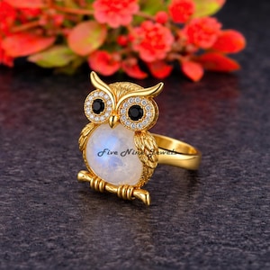 Peut inclure: Une bague dorée représentant un hibou avec un grand ventre en pierre de lune ronde et irisée, des yeux noirs circulaires et de petites pierres blanches entourant les yeux. Le texte Five Nine Jewels est visible sur le corps du hibou.