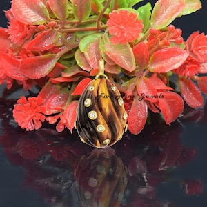 Puede incluir: Un colgante de oro en forma de huevo con un patrón de espiral marrón y amarillo. El huevo está decorado con piedras preciosas transparentes. El colgante cuelga de una cadena de oro.