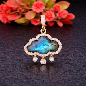 Puede incluir: Colgante dorado en forma de nube con una piedra central azul y verde. La nube está bordeada por pequeñas piedras transparentes, y tres piedras redondas y transparentes cuelgan debajo. El texto "Five Nine Jewels" es visible.