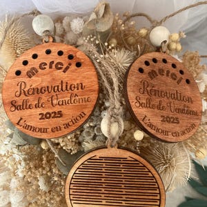Peut inclure: Trois ornements ronds en bois avec un design gravé au laser. Les ornements présentent le texte "Merci Rénovation Salle de Vendôme 2025 L'amour en action".