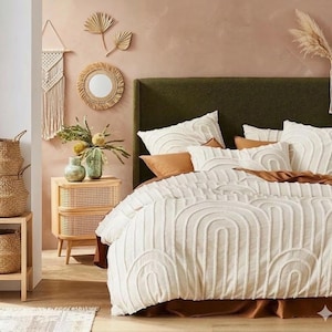 May include: A bedroom with a green headboard and a bed dressed with a white, arched-patterned duvet cover and pillows. The room includes a wooden bedside table with a woven front and a decorative mirror, creating a warm, inviting atmosphere.