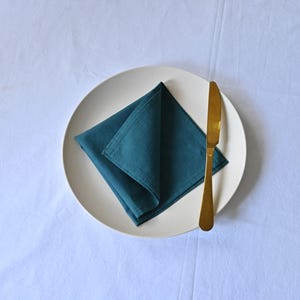 Könnte beinhalten: Ein teal-farbenes Leinen-Servietten auf einem weißen Teller, neben einem goldfarbenen Messer. Das Geschirr liegt auf einer weißen Tischdecke. Geeignet für ein Esszimmer- oder Küchenthema.