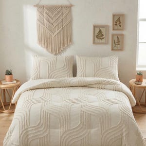 Könnte beinhalten: Ein cremefarbenes Bettwäsche-Set mit einem getufteten Wellenmuster, einschließlich einer Bettdecke und zwei Kissen. Ein Makramee-Wandbehang und gerahmte botanische Drucke schmücken die Wand über dem Bett. Zwei kleine Beistelltische mit Pflanzen befinden sich auf beiden Seiten des Bettes.