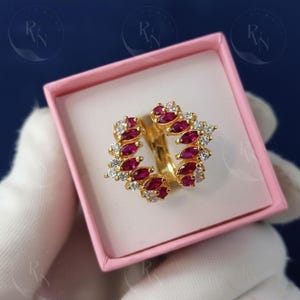 May include: A gold ring with a floral design, featuring alternating ruby and diamond-like stones. The ring is presented in a pink box, set against a dark blue background.