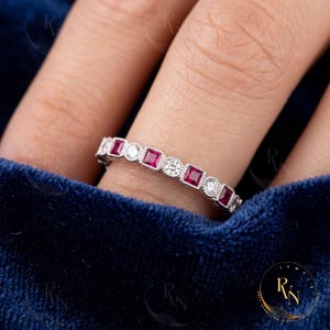May include: A silver-toned band ring featuring an alternating pattern of square-cut red rubies and round-cut white diamonds. The stones are set in a milgrain-detailed metal channel, creating a continuous, symmetrical design around the finger.