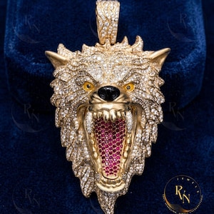 May include: A gold-colored pendant in the shape of a wolf's head, encrusted with clear gemstones. The wolf's mouth is open, revealing red gemstones. The eyes are yellow, and the nose is black. The pendant has a loop at the top for a chain.