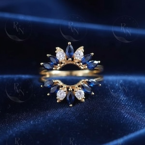 May include: A gold ring with a crown-like design, featuring marquise-cut blue gemstones and round white gemstones. The ring is set against a dark blue background, creating a striking contrast. The ring's design is elegant and eye-catching.