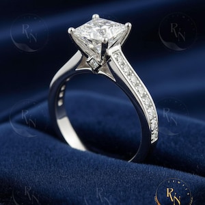 May include: A silver engagement ring with a large, square-cut diamond center stone. The band is adorned with smaller diamonds, and the ring is set against a dark blue background. The ring is a classic design.