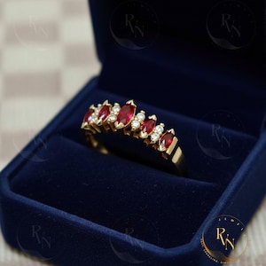 May include: A gold ring with a row of marquise-cut red gemstones and small, round, clear gemstones. The ring is set in a gold band and is displayed in a dark blue velvet ring box.