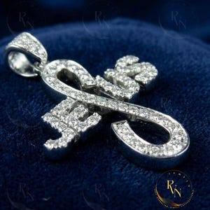 May include: A silver pendant with the letters "RN" encrusted with small, sparkling diamonds. The pendant is displayed on a dark blue velvet surface, with a logo in the bottom right corner.