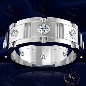 May include: A silver-colored ring with multiple clear, sparkling gemstones. The ring has a wide band with a series of vertical bars and a row of smaller stones set into the band. The design is symmetrical and modern, with a polished finish.