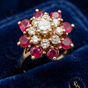 May include: A gold ring featuring a floral cluster design with a central round diamond surrounded by a layer of smaller round diamonds and an outer layer of vibrant red round rubies, all set in a multi-tiered arrangement on a gold band.