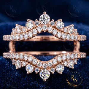 May include: A rose gold ring guard featuring two curved bands connected by a bridge. The bands are encrusted with numerous round, clear, brilliant-cut stones arranged in a crown-like pattern, creating a shimmering, multi-layered appearance.