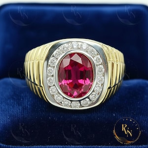 May include: A gold and silver ring with a large oval ruby gemstone at the center, surrounded by a halo of small diamonds. The ring has a ribbed design on the sides and is set against a blue velvet background.