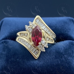 May include: A gold ring featuring a marquise-cut ruby center stone, surrounded by baguette and round-cut diamonds. The ring has a unique, asymmetrical design with a split shank and a cluster of diamonds on each side.