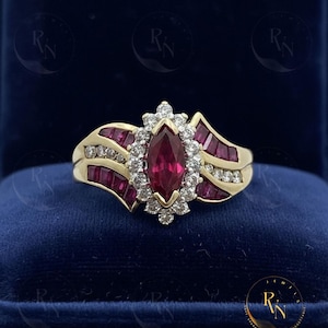 May include: A gold ring with a marquise-cut ruby center stone, surrounded by a halo of diamonds. The ring features additional ruby and diamond accents on the band, set against a dark blue velvet background. The ring is displayed in a jewelry box.