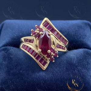 May include: A gold ring featuring a central marquise-cut ruby surrounded by smaller round rubies and two diagonal rows of channel-set baguette rubies. The design incorporates clusters of round rubies on the sides of the central stone.