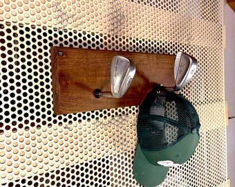 May include: A wall-mounted rack made from a rectangular wooden board featuring two silver metal golf club heads as hooks. A dark green baseball cap with a mesh back is hanging from the right golf club hook, displaying a white letter on the front.
