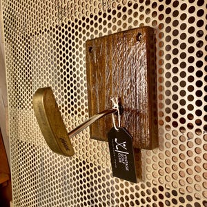 May include: A golf club head, gold in colour, mounted on a wooden plaque. The plaque is stained a dark brown. A black tag with "Vintage Golf Club" is attached. The background is a perforated metal sheet with small holes.