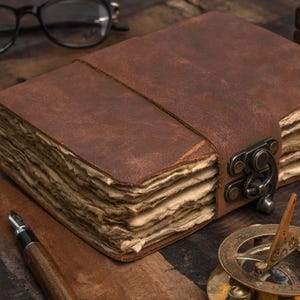 Puede incluir: Un diario de cuero marrón con cierre de hebilla y correa. El diario tiene páginas gruesas y envejecidas. Un bolígrafo, gafas y un reloj de sol también son visibles en la superficie de madera.