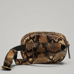 May include: A waist bag featuring a brown and black snakeskin pattern. The bag has a rounded rectangular shape with a matching snakeskin strap, a black interior lining visible at the edges, and a gold-toned metal buckle on the adjustable strap.