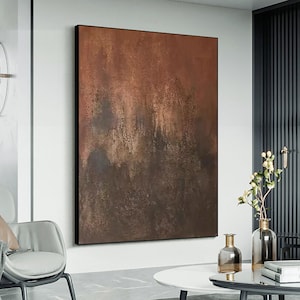 May include: Abstract painting with brown and gold hues, featuring a textured, layered effect. The painting is framed in a black frame and hangs on a white wall.