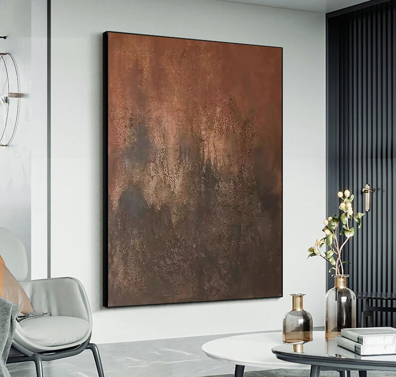 May include: Abstract painting with brown and gold hues, featuring a textured, layered effect. The painting is framed in a black frame and hangs on a white wall.