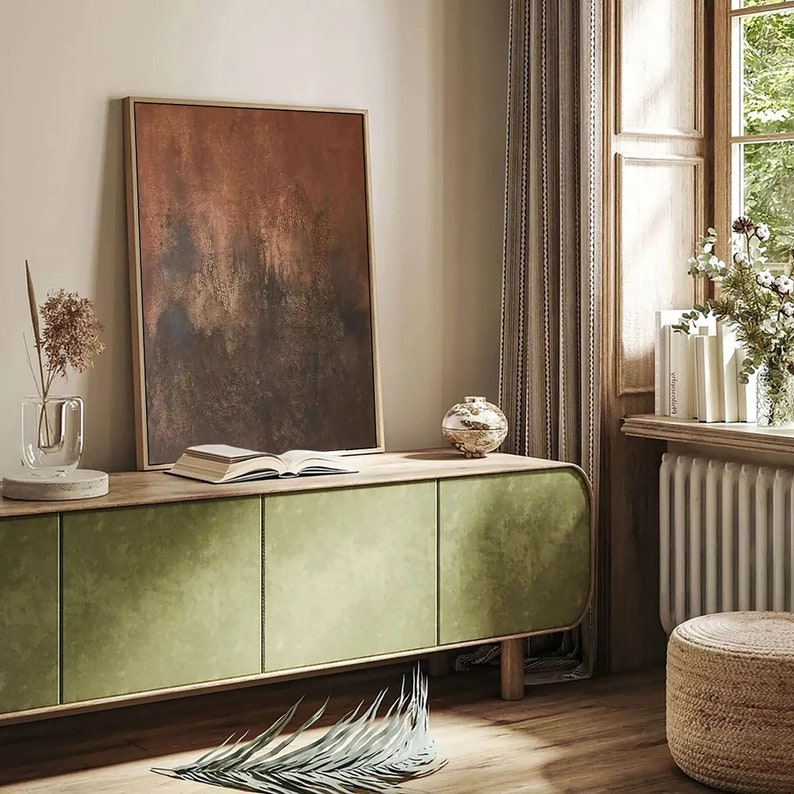May include: A green cabinet with three doors and rounded edges sits on a wooden floor. A book is on top of the cabinet, and a gold decorative object is on the right side. A painting with brown and orange hues hangs on the wall behind the cabinet.