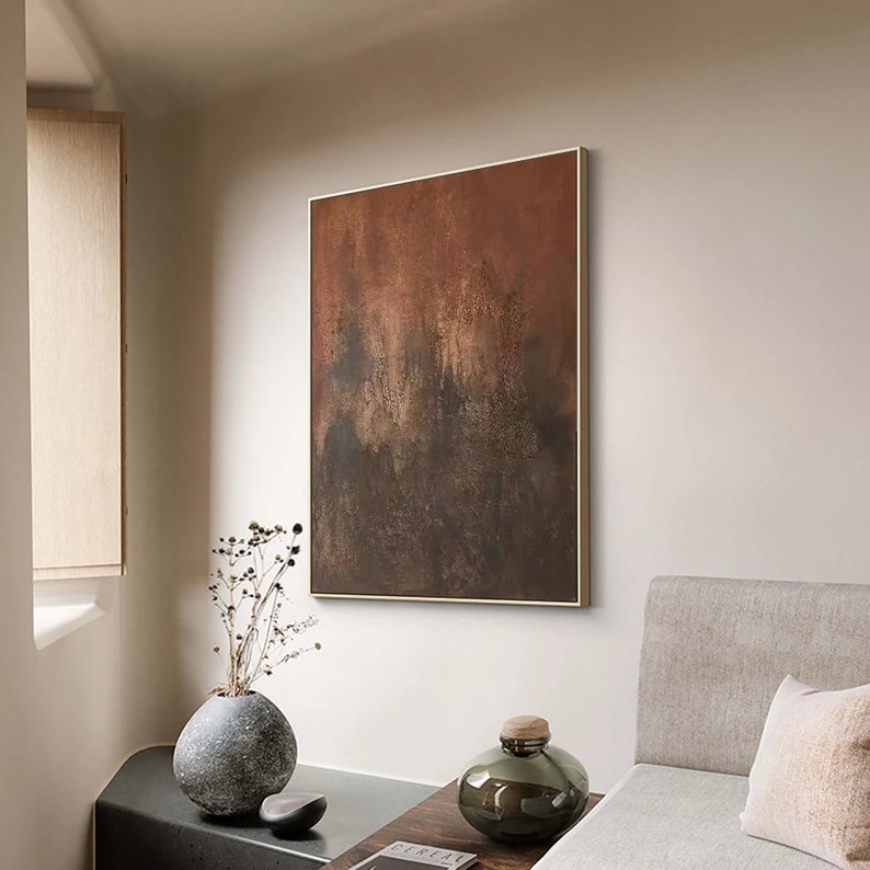 May include: A framed abstract painting with a brown, orange, and black colour scheme hangs on a white wall. A vase with dried flowers sits on a table in front of the painting.