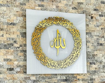 Op de afbeelding: Een gouden islamitisch kalligrafiekunstwerk met het woord "Allah" in het midden, omringd door een cirkel van Arabisch schrift. De kunst is gemonteerd op een witte achtergrond en ingelijst.