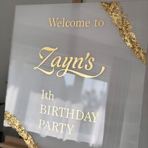 May include: A clear acrylic sign with gold lettering that reads "Welcome to Zayn's 1th BIRTHDAY PARTY." Gold leaf accents the corners. The sign is displayed on a wooden easel.