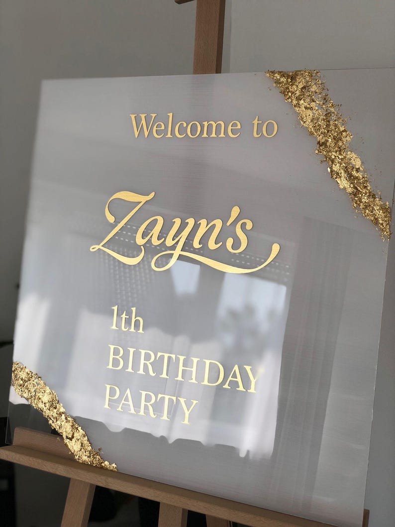 May include: A clear acrylic sign with gold lettering that reads "Welcome to Zayn's 1th BIRTHDAY PARTY." Gold leaf accents the corners. The sign is displayed on a wooden easel.